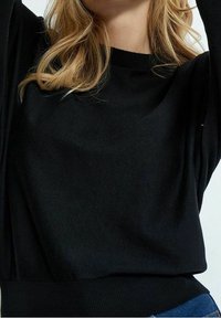 Mustahdin musta neulepaita, jossa on pyöreä kaula-aukko ja pitkät hihat. Pehmeä kangas, jossa on hienovarainen tekstuuri. Vyötäröltä istuva, minimalistinen muotoilu.