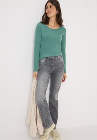 Langsleeve, teal top gecombineerd met vervaagde grijze jeans. Het model houdt een crèmekleurige fleecejas vast en draagt lichte groene sneakers.