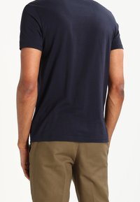 Mand iført en ensfarvet navy blå kortærmet T-shirt og lysebrune bukser, stående med ryggen mod kameraet.