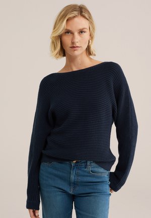 Blond kvinde iført en marineblå striktrøje og blå jeans, stående mod en ensfarvet beige baggrund, kiggende direkte ind i kameraet.