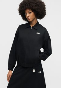 Femme aux cheveux afro portant une veste noire zippée et une jupe, toutes deux avec le logo The North Face, debout devant un fond clair uni.