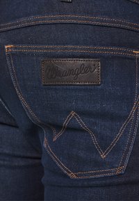 Jeans de mezclilla azul oscuro con una etiqueta de cuero Wrangler, costuras naranjas y un diseño 'W' característico en los bolsillos traseros.