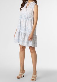 Mouwloze witte jurk met lichtblauwe geometrische patronen, gelaagd ontwerp en verstelbare strik bij de hals. Gecombineerd met beige hoge hakken.