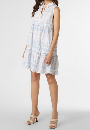 Mouwloze witte jurk met lichtblauwe geometrische patronen, gelaagd ontwerp en verstelbare strik bij de hals. Gecombineerd met beige hoge hakken.