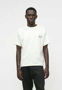 Licht crèmekleurig katoenen t-shirt met een ontspannen pasvorm, korte mouwen en een groene logo-print op de linkerkant van de borst. Gecombineerd met donkere jeans.