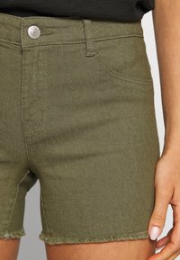 Olive green denim shorts with a frayed hem, featuring a front button closure and side pockets. Soft, textured fabric with a fitted design.