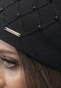 Zwarte gebreide beanie met een diamanten patroon, geaccentueerd door zwarte kralen en een metalen logo label. Close-upweergave benadrukt de textuur en details.