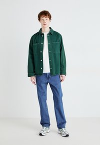 Zelena jeans jakna z dvema sprednjima žepoma, pod njo bela majica, modri jeans in sivi športni čevlji z zelenimi akcenti.