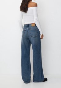 Kvinna klädd i högmidjade, vida blå jeans och en vit avskuren långärmad topp, stående mot en enkel vit bakgrund.