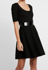 Svart klänning med en figursydd bodice, korta ärmar och en utsvängd kjol. Har ett glansigt svart lackbälte i midjan. Slät tygstruktur.