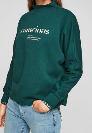 Grüner Pullover mit hohem Kragen, der ein grafisches Textdesign zeigt. Lange Ärmel mit floralen Akzenten an den Bündchen. Weiche Textur.
