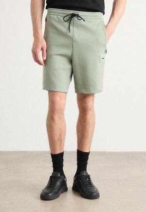 Light green shorts made of soft fabric, featuring a drawstring waistband, side pocket, and a minimalist design. Worn with black sneakers and socks.