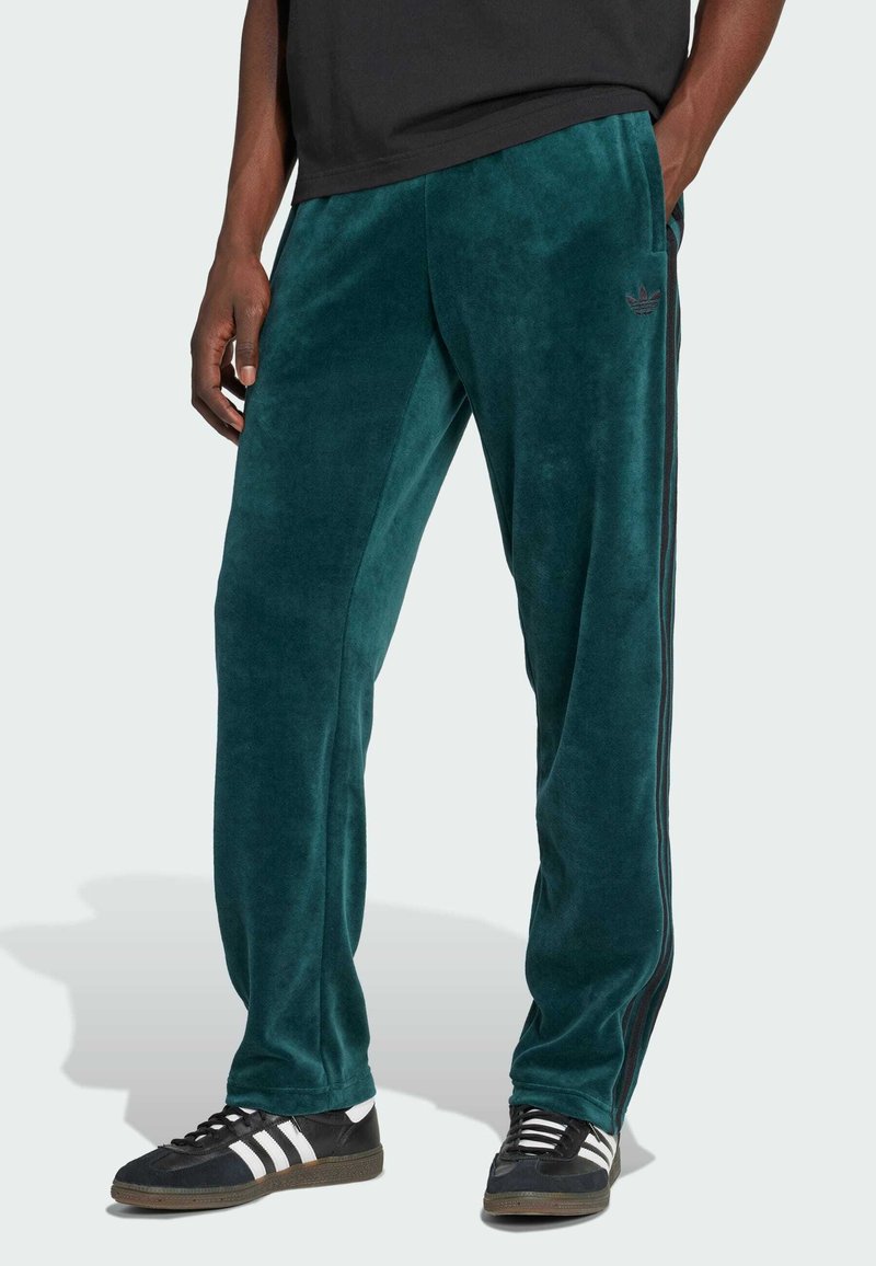 Hombre que lleva pantalones de chándal de terciopelo color teal con franjas laterales y zapatillas negras con franjas blancas, de pie con una mano en el bolsillo.