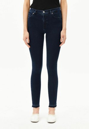 Vrouw draagt donkerblauwe skinny jeans met rafelige zoom en witte ballerina's, staand tegen een effen witte achtergrond.