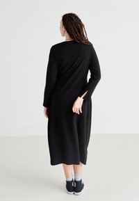 Robe noire à manches longues avec une coupe ample, longueur genoux et fentes latérales. Associée à des baskets noires et des chaussettes gris clair.