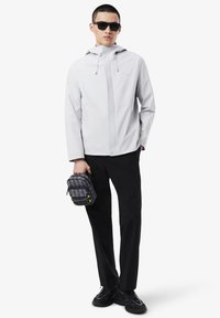 Chaqueta impermeable gris claro con capucha, pantalones negros y zapatillas negras. Sosteniendo una pequeña bolsa negra con estampado y detalles amarillos.