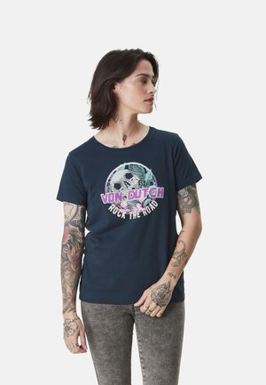 Personne aux cheveux foncés et tatouages portant un t-shirt bleu marine avec un motif de crâne et de fleurs ainsi que le texte "Von Dutch Rock the Road", regardant sur le côté.