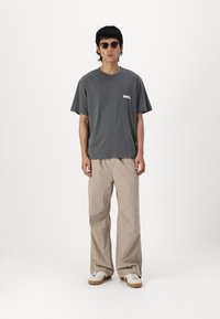 Camiseta de algodón gris con un logo en el lado izquierdo del pecho, combinada con pantalones beiges holgados y zapatillas blancas con suelas marrones. Gafas de sol marrones puestas.