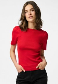 Maglietta a coste rossa con maniche a sbuffo corte, scollatura rotonda e silhouette aderente. Abbinata a pantaloni neri.