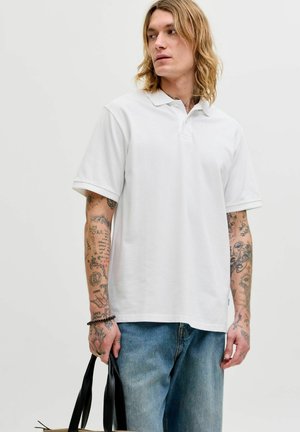 Ung mann med langt hår iført hvit polotrøye og blå jeans, med tatoverte armer, holder en svart skinnveske i venstre hånd.