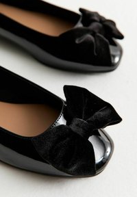 Bailarinas de charol negro con grandes lazos de terciopelo negro en la punta, mostradas sobre un fondo blanco.