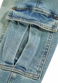 Tela de denim azul claro con un bolsillo con solapa, costuras visibles y un leve desvanecimiento. Muestra textura y variaciones de color distintas.