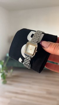 Sudraba nerūsējošā tērauda pulkstenis ar taisnstūrveida seju, zelta akcentiem un saistītu aproci, novietots uz melnā spilvena.