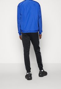 Modrá nylonová bunda s límcem do stoje, doplněná černými tepláky se světlými pruhy po stranách a elastickými manžetami, nošená s černými sportovními botami.