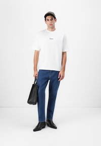 Hvit kortermet t-skjorte med "Selected"-logo, blå denimjeans, svarte lærstøvler og en svart bag. Modellen står mot en hvit bakgrunn.