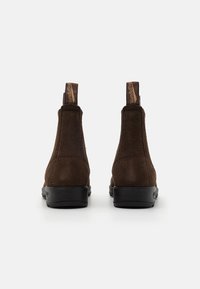 Botines de ante marrón con suela negra lisa y lengüetas para facilitar su colocación. El diseño presenta una punta redondeada y detalles de costura minimalistas.
