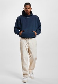 Sudadera con capucha azul marino y bolsillo canguro frontal, combinada con pantalones beige de corte relajado y zapatillas blancas. Textura suave.