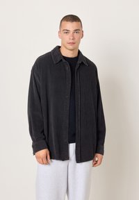 Mladić s kratkom kosom koji nosi crnu kordiranu košulju s gumbima preko crne majice i svijetlosivih hlača, stojeći pred jednobojnom pozadinom.