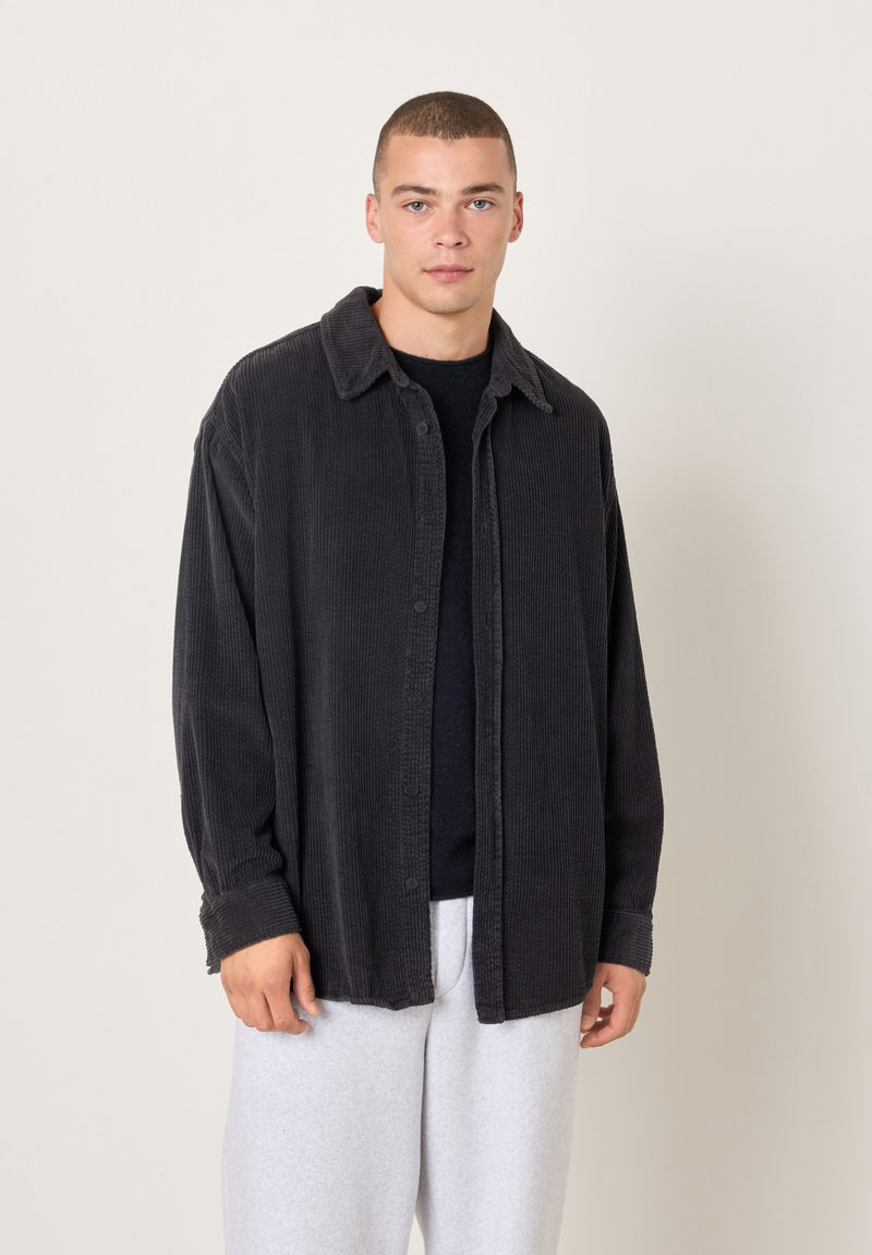 Mladić s kratkom kosom koji nosi crnu kordiranu košulju s gumbima preko crne majice i svijetlosivih hlača, stojeći pred jednobojnom pozadinom.