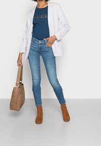 Blazer blanco, camiseta navy con el logo "GUESS", jeans skinny azules, botas tobilleras marrones y un bolso de ante color marrón claro con una correa.