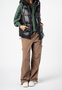 Schwarze wattierte Weste mit Kapuze über einem grünen Pullover, kombiniert mit beigefarbenen Cargohosen und weißen Turnschuhen. Die Weste verfügt über Reißverschlusstaschen.