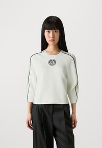 Maglione bianco a coste con maniche a tre quarti, caratterizzato da un bordo nero lungo le spalle e un patch centrale con logo.