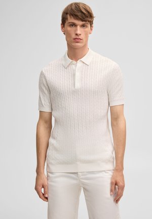 Młody mężczyzna w białym polo z krótkim rękawem o warkoczowym splocie i białych spodniach, stojący na jednolitym szarym tle.