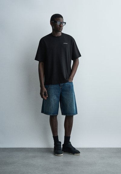 Άνδρας που στέκεται μπροστά σε γκρι τοίχο, φορώντας μαύρο υπερμέγεθος μπλουζάκι, τζιν σορτς μέχρι τα γόνατα, μαύρες κάλτσες, μαύρα αθλητικά παπούτσια και σκούρα γυαλιά ηλίου.