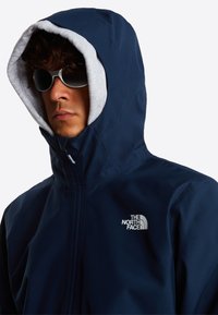 Veste à capuche bleu marine avec doublure en polaire grise, fermeture éclair, et logo The North Face sur la poitrine. Le mannequin porte des lunettes de soleil élégantes.