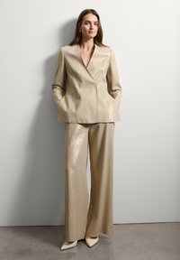 Blazer metálico beige con solapa muestrera, combinado con pantalones de pierna ancha a juego. Textura suave, diseño minimalista y tacones blancos de punta.