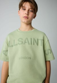 Lys grøn t-shirt i bomuld med korte ærmer. Har præget tekst "ALLSAINTS LONDON" på forsiden. Tekst og stof har en glat overflade.