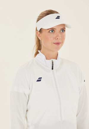 Kvinde med blondt hår bundet tilbage iført en hvid sportskasket og en hvid lynlåsjakke med et lille navy-logo på brystet.