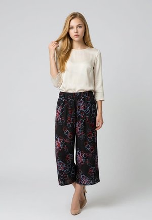 Joven mujer con cabello largo y rubio lleva una blusa crema y pantalones anchos negros con patrones geométricos rojos y azules, de pie frente a un fondo liso.