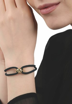 Nahaufnahme eines Handgelenks mit einem schwarzen, gedrehten Seilarmband mit goldenem Verschluss, neben einem Gesicht mit neutralem Ausdruck und sanft rosa Lippen.