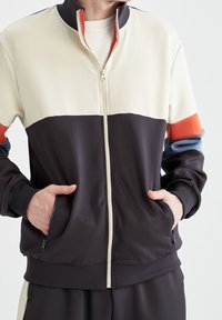 La veste en coloris bloc présente un haut crème, un bas bleu marine foncé et des accents rouges et bleus, avec une fermeture éclair, des poches et des poignets côtelés.