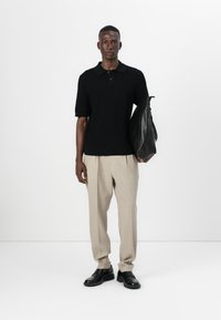 Polo shirt noir à côtes avec des manches courtes, associé à un pantalon plissé beige clair et des chaussures en cuir noires. Tenant un sac en cuir noir.