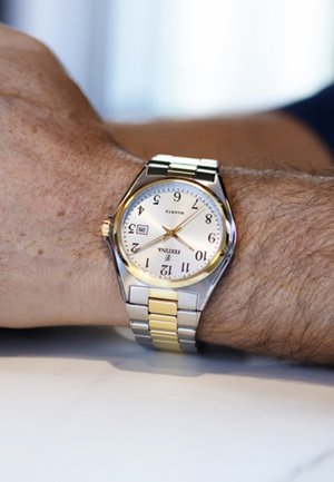 Montre en argent et or avec un cadran rond, un fond crème, des chiffres noirs et un affichage de la date. Bracelet en maillons métalliques avec des accents polis.