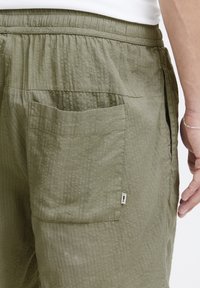 Shorts vert olive en tissu texturé, dotés d'une taille élastique et d'une seule poche latérale avec une petite étiquette de logo.