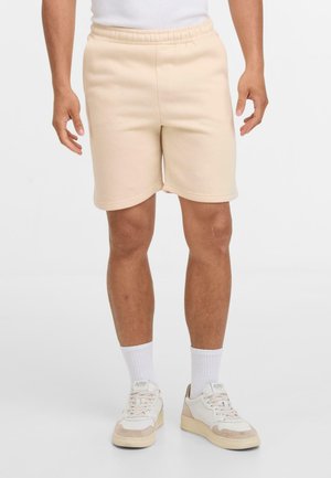Shorts en coton beige clair avec une taille élastique, des poches latérales et une texture lisse, assortis à des baskets blanches et des chaussettes mi-mollet.