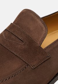Mocassino in suede marrone con una fascia rialzata sulla parte superiore e dettagli di cucitura sottili. La fodera interna è di un colore tan a contrasto.